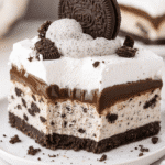 Oreo Ice Cream Cake 3 A close-up of a decadent Oreo ice cream lasagna slice on a white plate, layered with a chocolate cookie crust, cookies and cream ice cream, chocolate pudding, whipped cream, and topped with a whole Oreo and crumbled cookie pieces.